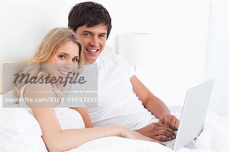 A man and woman with a laptop on the bed smile as they look in front of them.