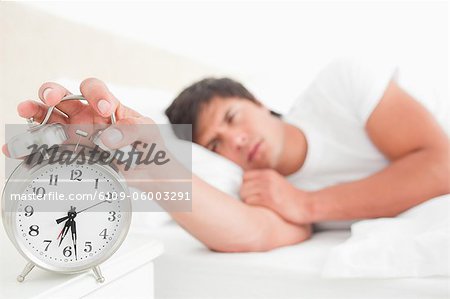 Close up and focus on the alarm clock which the half awake man is turning off with his hand.
