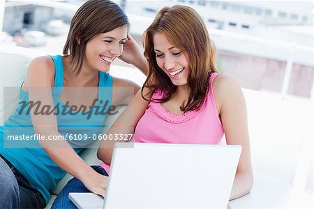 Young woman holding a laptop accompanied by her friend sitting on a white sofa