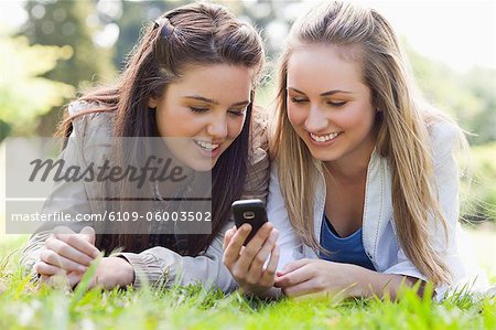 Two smiling friends lying on the grass in a park while looking at a text on a cellphone