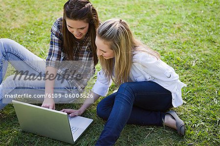 Young pretty girls sitting down on the grass in a park while looking at a laptop