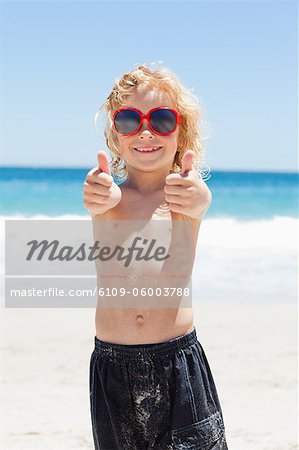 Smiling little boy with sunglasses at the beach giving thumbs up