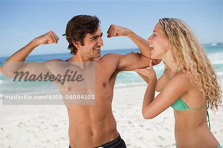 Young smiling woman holding her boyfriend's biceps while standing on the beach