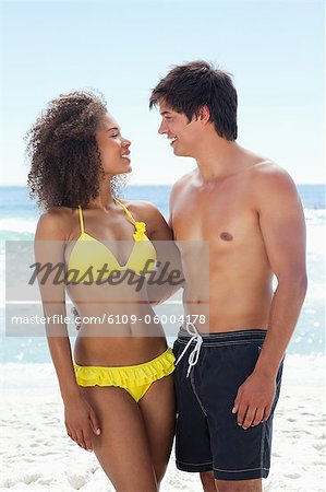 A man and a woman wearing swimsuits smiling as they look at each other while standing on a beach