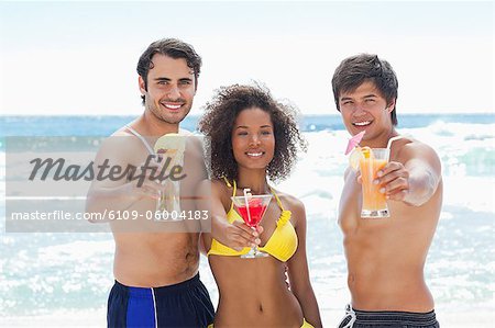 Two men and a woman in swimsuits smiling as they offer cocktails on a beach