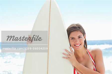 Woman in bikini smiling as she holds a surfboard closely with two hands on the beach
