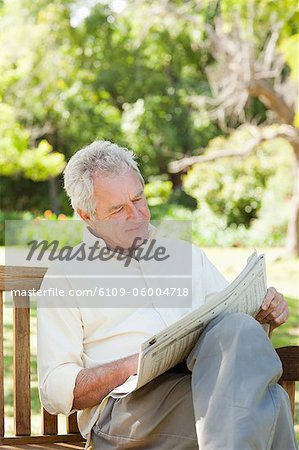 Man looking at a newspaper while sitting on a wooden bench in a park