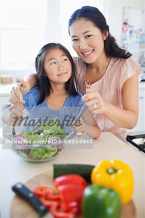A mother and daughter tosses a salad while the mother looks ahead and the daughter looks up at her mom