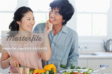 A husband and wife work and eat in the kitchen making a salad and eating pepper