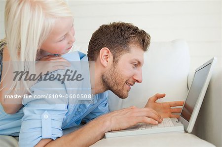 A smiling father lies on the couch as his daughter lies on top of him, while he uses his laptop