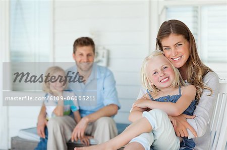 A smiling mother and daughter embrace as the husband and son embrace while sitting behind them