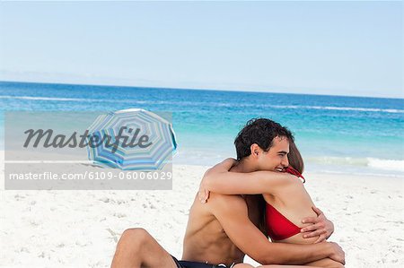 couple making a hug sitting on the beach with a parasol and the sea in background