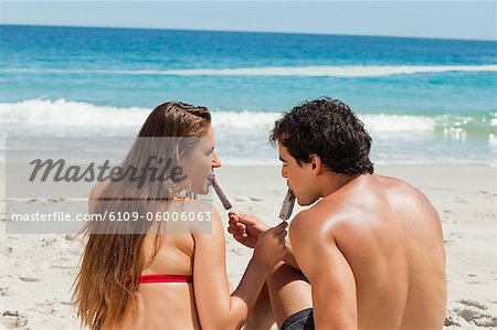 Rear view of a tanned couple eating Popsicle together in front of the sea