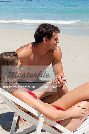 Woman on a deck chair while her attractive boyfriend holds her hand with sea in background