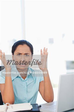 Upset secretary raising her hands while sitting at a desk in front of her laptop
