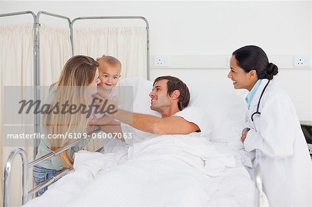 Relaxed man lying in a hospital bed while being given his smiling baby by his wife Relaxed man lying in a hospital bed while being given his smiling baby by his wife