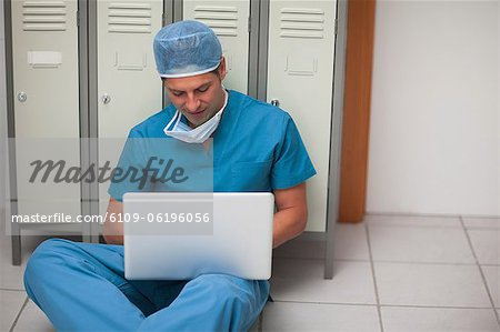 Male surgeon sitting on the floor of a locker while holding a laptop
