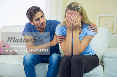 Crying blonde woman sitting on a couch being consoled by her boyfriend