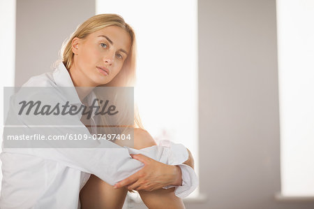 Pretty blonde wearing mens shirt sitting on bed pouting at camera at home in bedroom