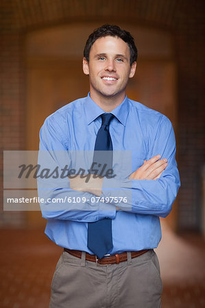 Cheerful handsome lecturer standing arms crossed in hallway in a college