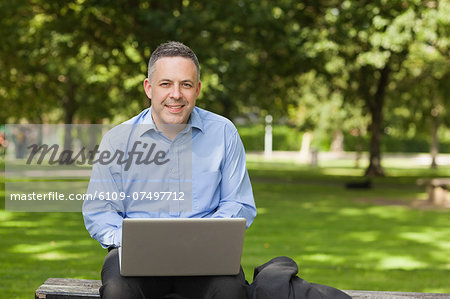 Cheerful professor sitting on bench using laptop on campus at the university