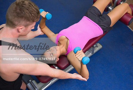 Trainer correcting toned brunette exercising with dumbbells in weights room of gym
