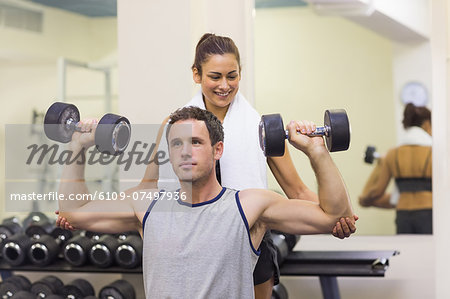 Trainer correcting muscular man lifting dumbbells in weights room of gym