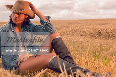 Beautiful young woman in hat sitting at cereal field against cloudy sky in summer