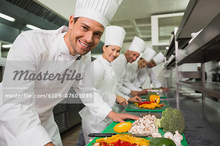 Row of trainee chefs slicing vegetables with one smiling at camera