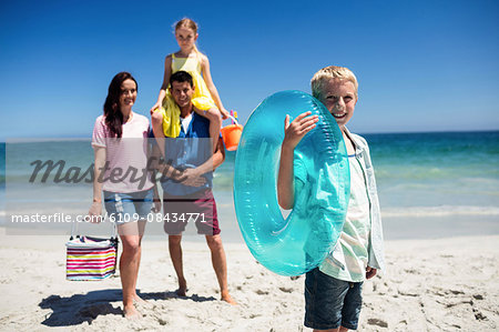 Cute boy holding inflatable buoy on the beach with family in background