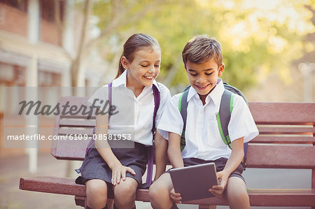 Happy school kids sitting on bench and using digital tablet