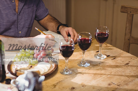 Cropped image of mature man writing in a book while tasting red wine and eating cheese