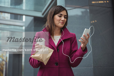 Businesswoman holding disposable coffee cup and parcel while listening to music