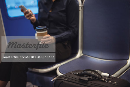 Businesswoman with coffee cup and luggage using phone while sitting in train