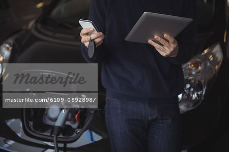 Mid section of man using digital tablet and mobile phone while charging electric car in garage