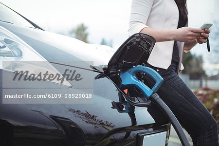 Mid section of woman using mobile pone while charging electric car on street