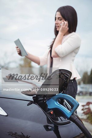 Beautiful woman talking on mobile phone while charging electric car on street