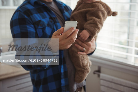 Mid section of father using mobile phone while holding his baby in kitchen