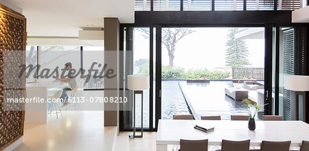 Young man using laptop in modern kitchen, dining area with patio door and swimming pool in foreground