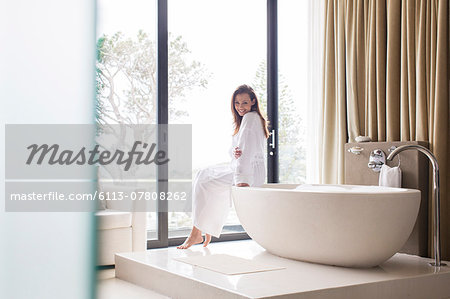 Portrait of smiling woman wearing white bathrobe, sitting on edge of bathtub in bathroom