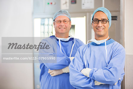 Two smiling doctors with arms crossed, wearing surgical clothing and eyeglasses in hospital