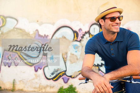 Smiling man in hat and sunglasses leaning on bicycle next to urban graffiti wall