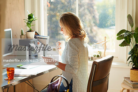 Woman drinking coffee and reading paperwork at desk in sunny home office