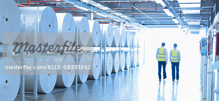 Workers in reflective clothing walking along large paper spools in printing plant