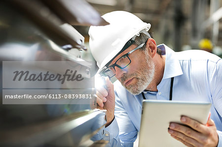 Focused engineer with digital tablet and flashlight examining part in factory