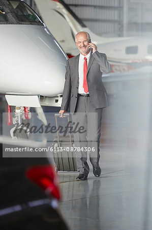 Smiling businessman pulling suitcase talking on cell phone in airplane hangar