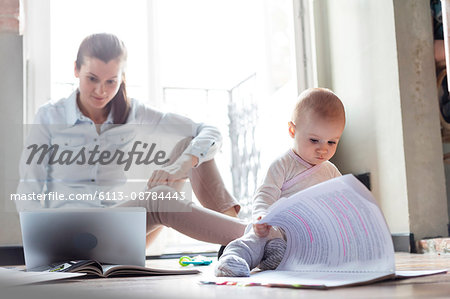 Curious baby daughter looking at paperwork next to mother working at laptop