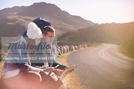 Young couple with backpacks hiking, resting at sunny, remote roadside with map and smart phone Young couple with backpacks hiking, resting at sunny, remote roadside with map and smart phone