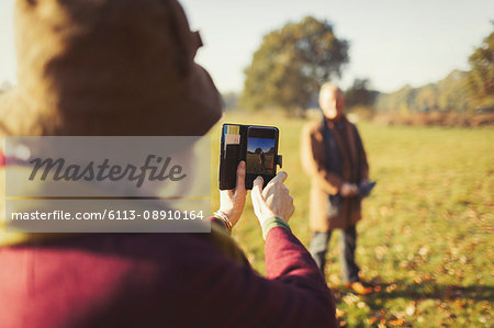 Senior woman with camera phone photographing husband in sunny autumn park