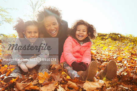 Portrait playful mother and daughters in autumn leaves in sunny park